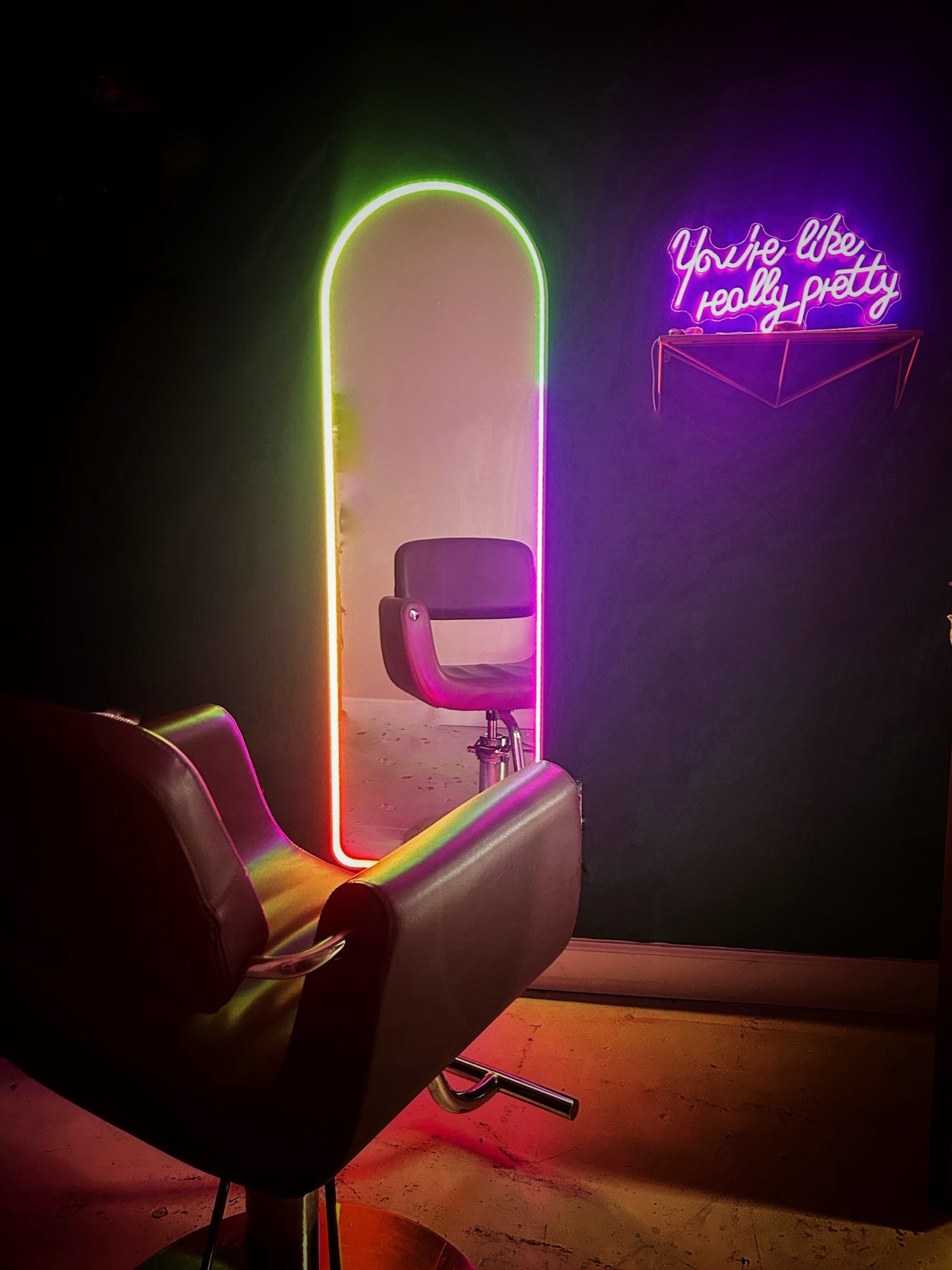 The styling chair by an arched mirror rimmed in rainbow light, beside a pink neon sign.