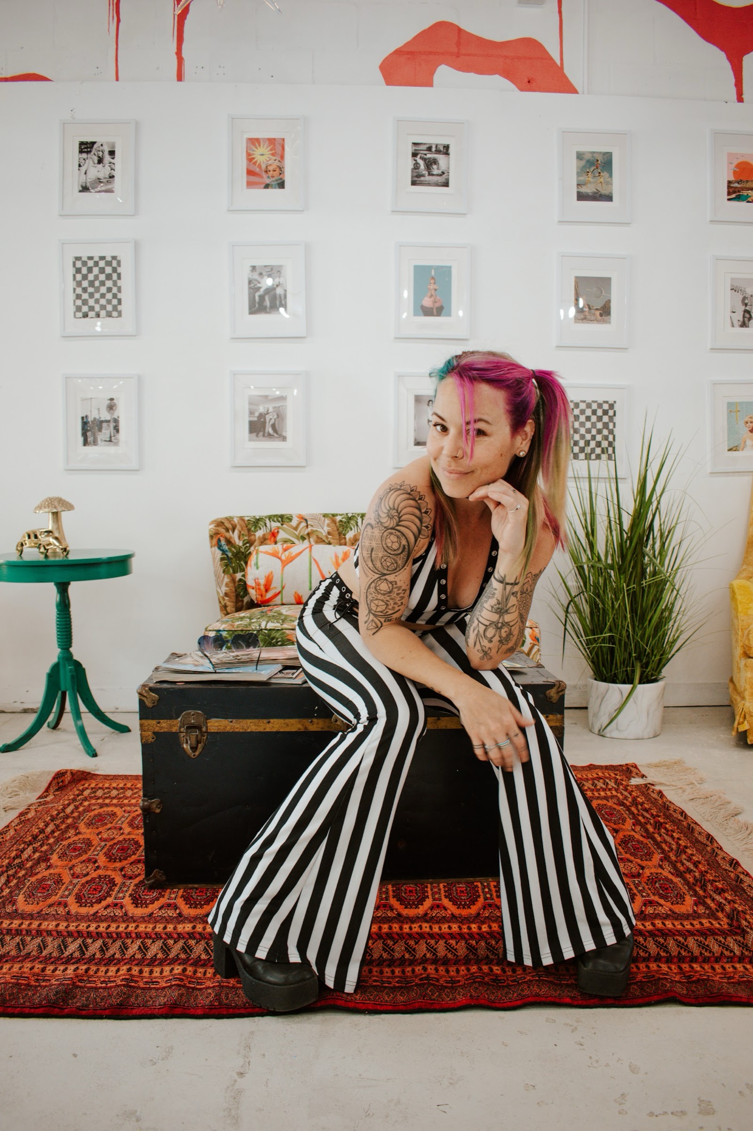 Shannon, in a black and white striped jumpsuit with pink hair, seated in her bright art-filled studio.